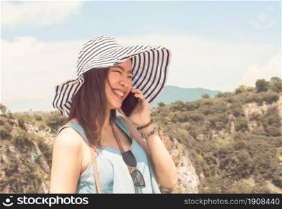 Asian beautiful woman wearing hat and talking on mobile phone while traveling for summer trip with blur background of mountains