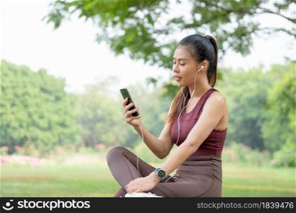Asian beautiful woman with sportwear sit and relax also enjoy her mobile phone in park or garden in morning during exercise.