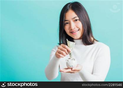 Asian beautiful young woman holding piggy bank on hands and putting dollars money banknotes, portrait of happy female smiling hold piggybank in palm, isolated on blue background, saving money concept