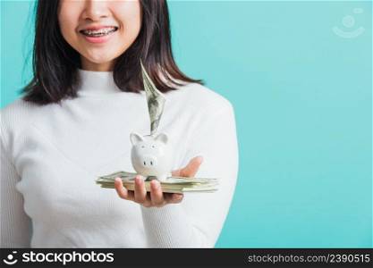 Asian beautiful young woman holding piggy bank on hands and putting dollars money banknotes, portrait of happy female smiling hold piggybank in palm, isolated on blue background, saving money concept