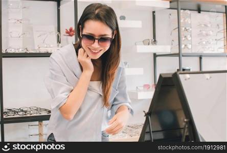Asian beautiful young woman smiling, looking at mirror, wearing and trying eyesight glasses in store. Sales, Discount and Optical Concept.