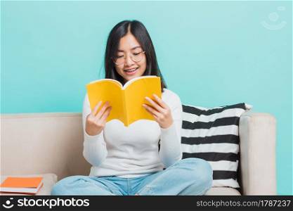 Asian beautiful young woman wearing eyeglasses sitting on sofa reading storybook, portrait relaxation of happy female smiling in living read a book studio shot isolated on blue background