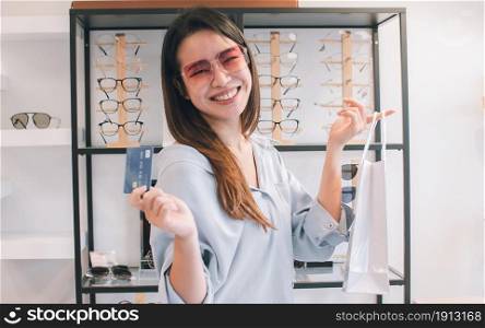 Asian beautiful young woman wearing fashion eyeglasses, holding credit card for shopping in glasses store and showing white bag. Sales, Discount and Optical Concept.