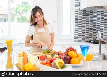 Asian beauty woman cooking and slicing vegetable in kitchen room with full of food and fruit on table for party. Holiday and Happiness concept. People and lifestyles concept. Girl looking at camera