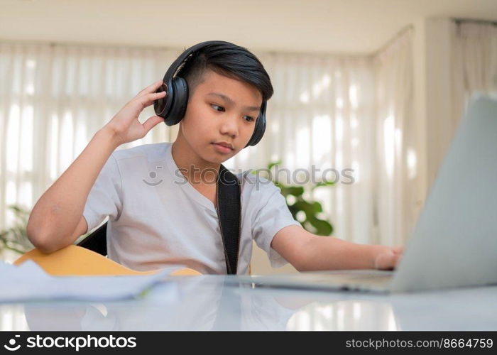 Asian boy playing guitar and watching online course on laptop while practicing for learning music or musical instrument online at home. Boy students study online with video call teachers play guitar.