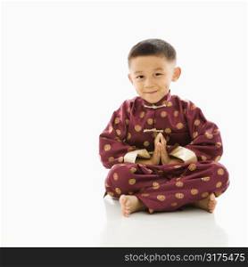 Asian boy sitting meditating against white background in traditional attire.