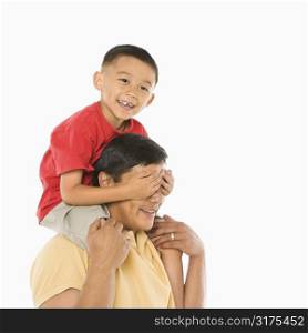 Asian boy sitting on father&acute;s shoulders with hands over his eyes in front of white background.