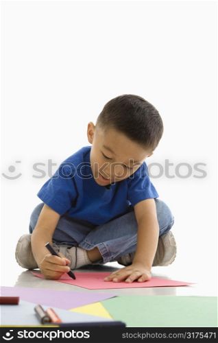 Asian boy sitting on floor coloring on paper.