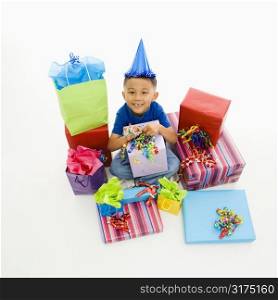 Asian boy wearing party hat sitting with pile of wrapped presents.