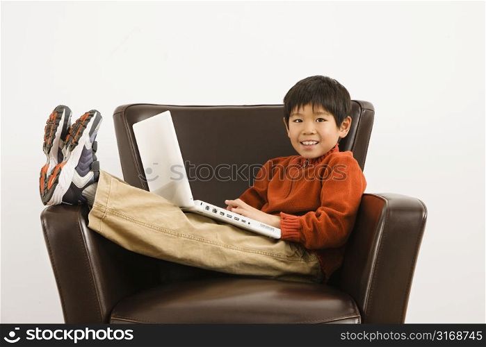 Asian boy with laptop computer sitting in chair smiling.