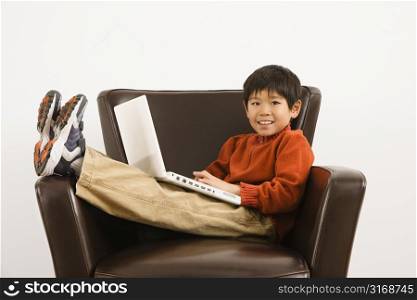 Asian boy with laptop computer sitting in chair smiling.