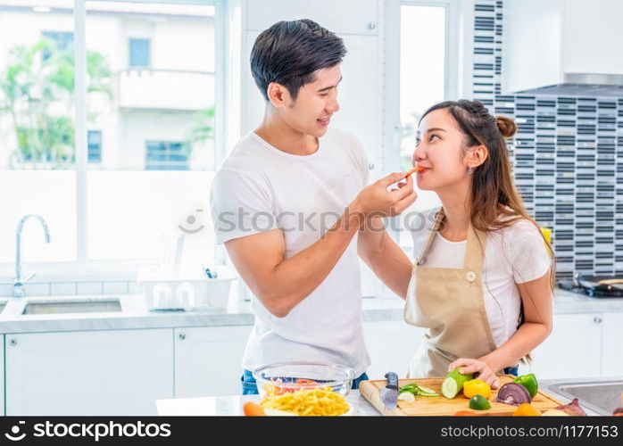 Asian boyfriend feeding fruit and vegetable to girlfriend to each other during preparing for dinner in home kitchen. Couple and Family concept. Honeymoon and Holidays. Man and woman cooking happily