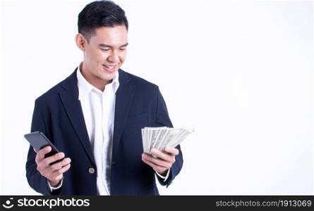 Asian business man wearing suit, using mobile phone and holding money with smiling happy face and standing on white background