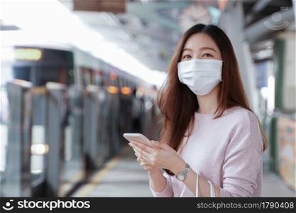Asian business woman in casual clothes wearing face mask using mobile phone. She is waiting for the train to go to work on the platform station. New normal lifestyle in city concept