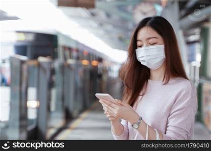 Asian business woman in casual clothes wearing face mask using mobile phone. She is waiting for the train to go to work on the platform station. New normal lifestyle in city concept