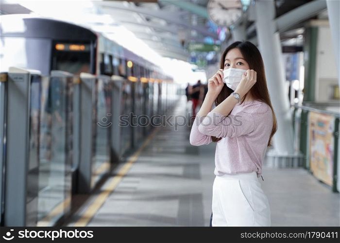 Asian business woman in casual dress code wearing face mask. She is waiting for the train to go to work on the platform station. New normal lifestyle in city concept