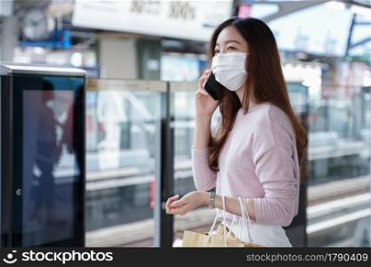 Asian business woman in casual dress code wearing face mask talking on mobile phone. She is waiting for the train to go to work on the platform station. New normal lifestyle in city concept