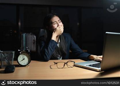 Asian business woman is sitting yawning with a hand covered her mouth, Laptop, on the desk, While the young woman works overtime in the office at night