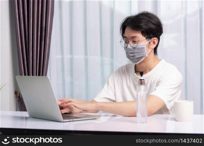Asian business young handsome man glasses, wearing protective mask with disinfectant gel beside he work from home office remote online with laptop computer quarantines disease coronavirus or COVID-19