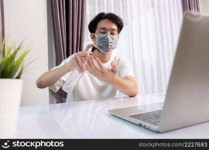 Asian business young handsome man glasses working from home office he quarantines disease coronavirus or COVID-19 wearing protective mask and cleaning hands with sanitizer gel on front laptop computer