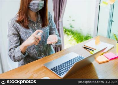 Asian Business young woman work from home office he quarantines disease coronavirus or COVID-19 wearing a protective mask and cleaning hands with sanitizer Spray on front laptop computer