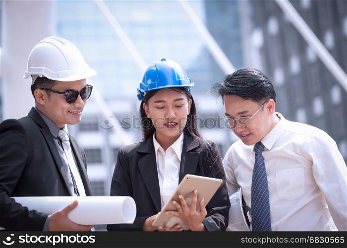 Asian businessman and woman discuss with engineer architect professional wearing safety helmet and suite, looking at tablet and holding construction roll plan for working team concept