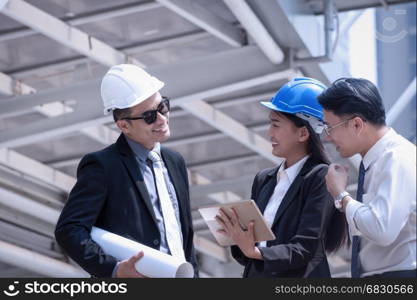 Asian businessman and woman discuss with engineer architect professional wearing safety helmet and suite, looking at tablet and holding construction roll plan for working team concept