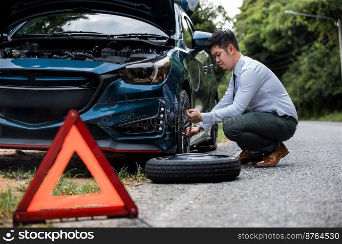 Asian businessman car broken has problems with wheel of his car during go to work in morning he changing replace rubber tire on wheels, business man have warning sign on road, problem transportation