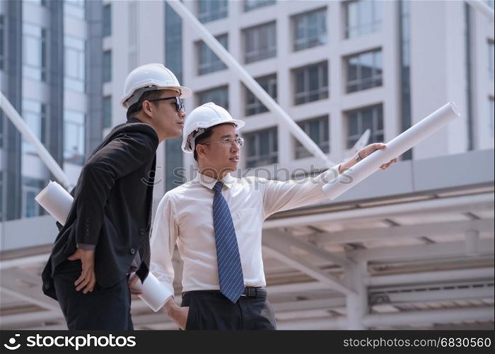 Asian businessman discuss with engineer architect professional wearing safety helmet and suite, looking and point to finger forward away and holding construction roll plan for working team concept