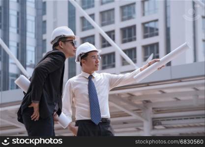 Asian businessman discuss with engineer architect professional wearing safety helmet and suite, looking and point to finger forward away and holding construction roll plan for working team concept