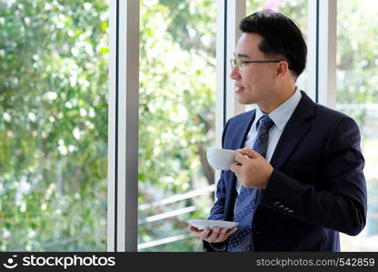 Asian Businessman holding coffee cup standing by windows, inside eco office building background, businessperson, office lifestyle