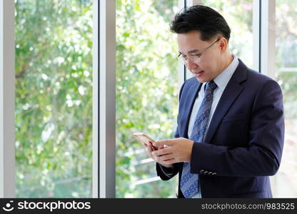 Asian Businessman holding smart phone with happy smiling face standing by windows, inside eco office building background, businessman on smart phone