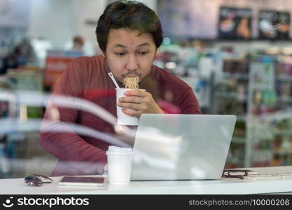 Asian businessman in casual suit eating noodles with urgent action in rush hour at the desk beside the glass in modern office, Business work hard concept