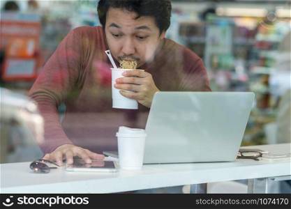Asian businessman in casual suit eating noodles with urgent action in rush hour at the desk beside the glass in modern office, Business work hard concept