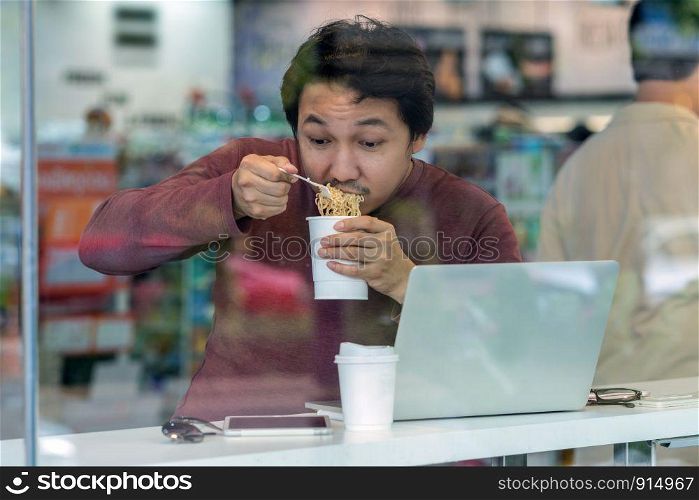 Asian businessman in casual suit eating noodles with urgent action in rush hour at the desk beside the glass in modern office, Business work hard concept