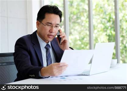 Asian businessman reading papers talking phone and working with laptop computer at office, business concept
