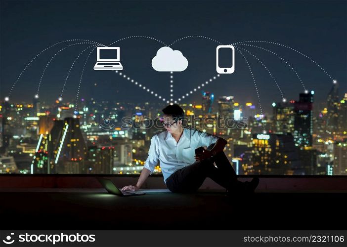 Asian businessman sitting and using the laptop with smart mobile phone for operate various icon of smart city Internet of Things Technology over the cityscape background, technology with IOT concept.