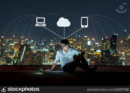 Asian businessman sitting and using the laptop with smart mobile phone for operate various icon of smart city Internet of Things Technology over the cityscape background, technology with IOT concept.