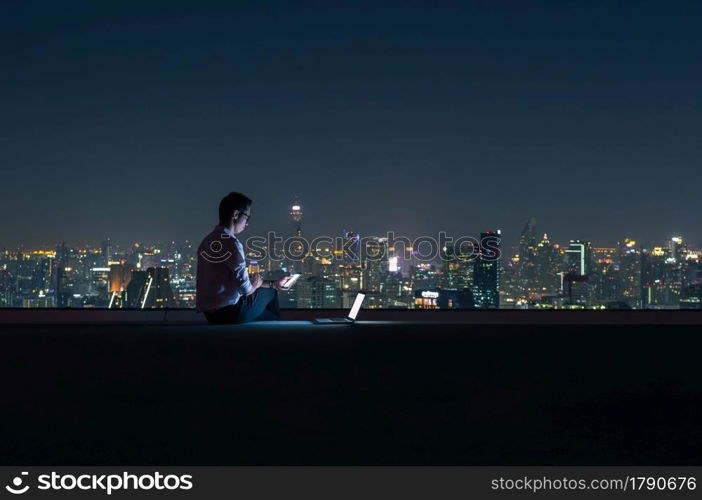 Asian businessman sitting at the top of the building. Use the smartphone and laptop computer with the scenery of the night view of the city, Business success and technology concept.. working late night