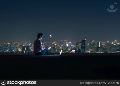 Asian businessman sitting at the top of the building. Use the smartphone and laptop computer with the scenery of the night view of the city, Business success and technology concept.. working late night