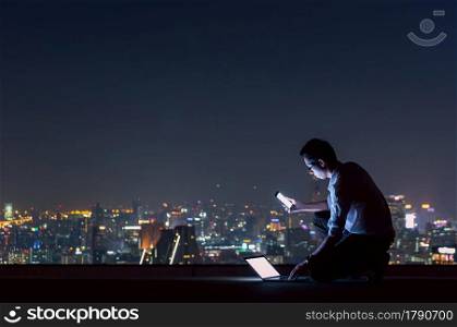 Asian businessman sitting at the top of the building. Use the smartphone and laptop computer with the scenery of the night view of the city, Business success and technology concept.. working late night