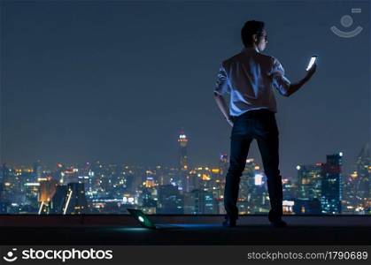 Asian businessman stand at the top of the building. Use the smartphone and laptop computer with the scenery of the night view of the city, Business success and technology concept.. working late night