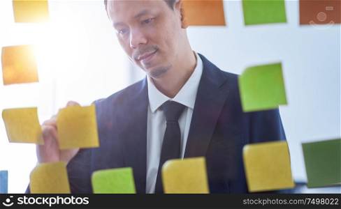 Asian businessman stand in glasses board with stickers and writing ideal , brainstorming concept .