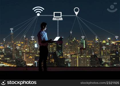 Asian businessman standing and using the laptop for operate various icon of smart city Internet of Things Technology over the cityscape background, technology with IOT concept.