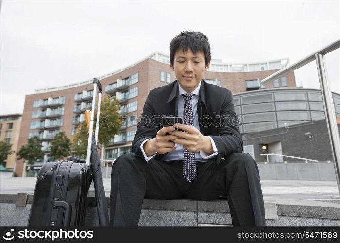 Asian businessman with luggage text messaging through cell phone against buildings