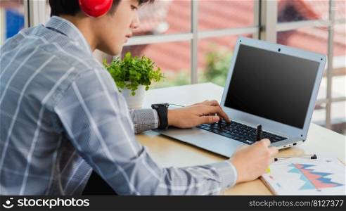 Asian businessman writing notes with pen inspired ideas list on paper notepad while working on laptop computer. Happy handsome man sitting busy on desk table working from home office doing note