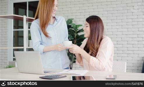 Asian businesswoman giving coffee to her colleague who is working with laptop at office. Group of casually dressed business people discussing ideas in the office.