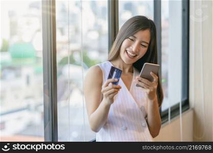 Asian businesswoman in casual suit doing online shopping by using credit card and paying via smart mobile phone application or technology laptop in modern office, business and lifestyle concept