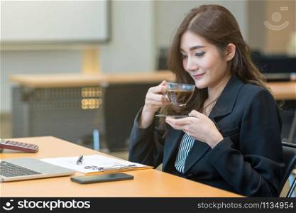 Asian Businesswoman in formal suit drinking coffee when working with computer laptop and office supplies in modern office or meeting room, Business and worker concept
