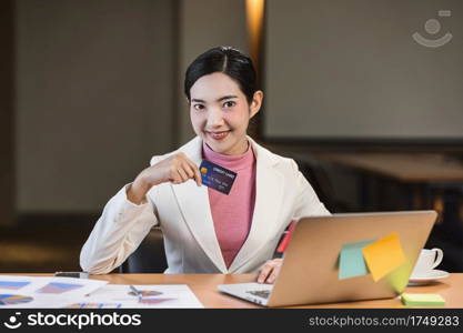 Asian businesswoman in formal suit holding and presenting credit card mockup for online shopping via technology laptop and mobile phone in modern office, business and lifestyle concept